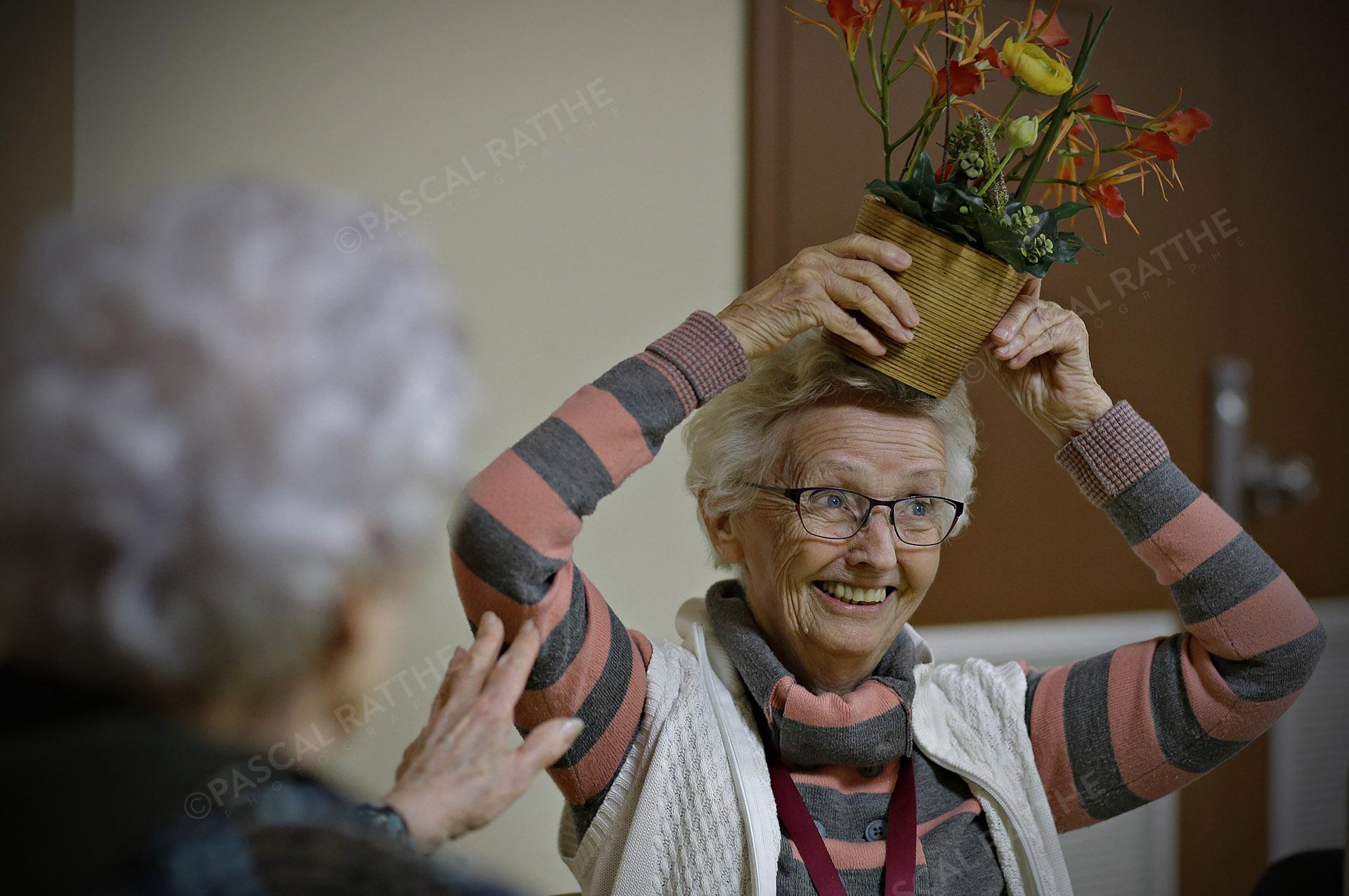 une dame du 3e âge avec un sourire éclantant et un pot de fleur sur la tête